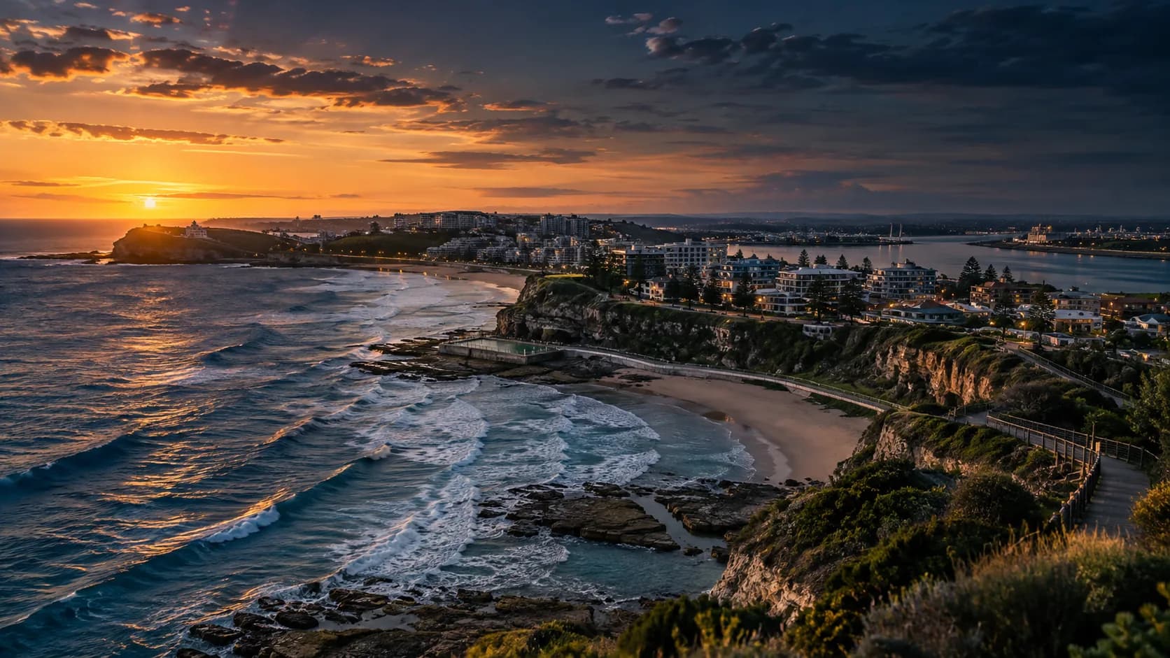 The Newcastle coastline at sunset, surf breaking below a headland of clifftop apartments and an ocean pool